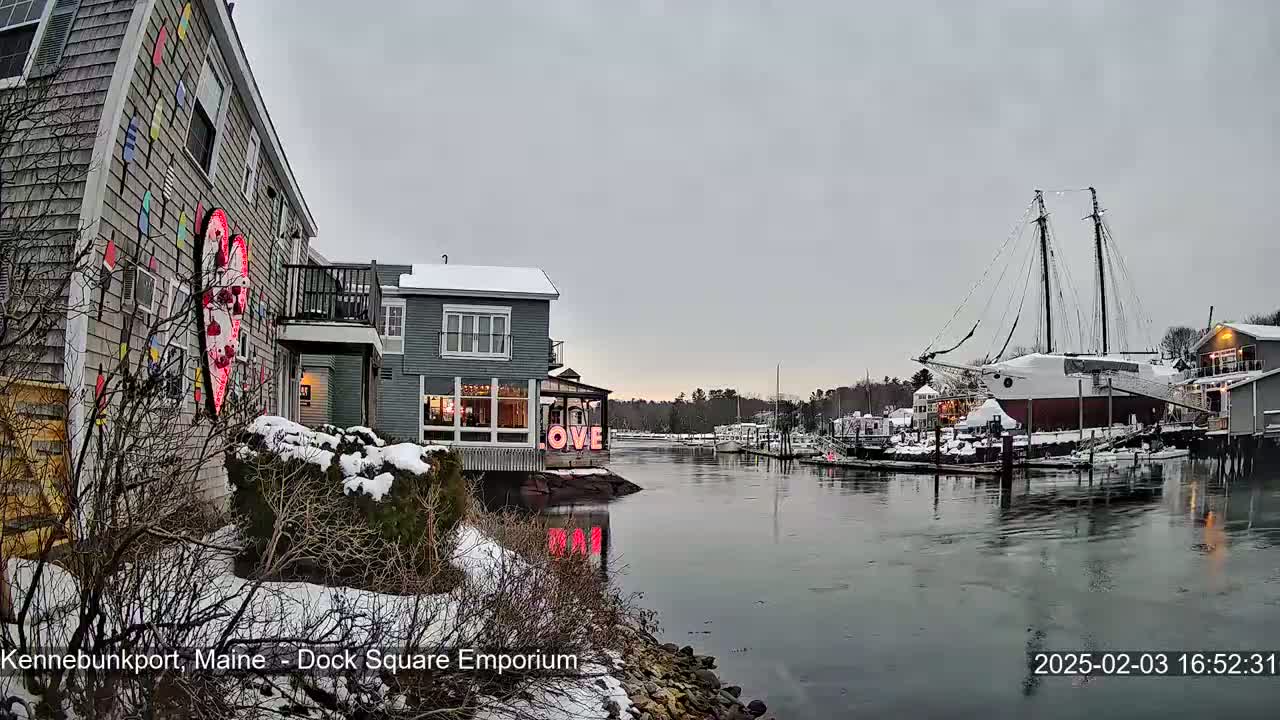 A snow-covered waterfront scene shows buildings with a large heart-shaped sign, and a large sailboat docked in a calm, overcast harbor.
