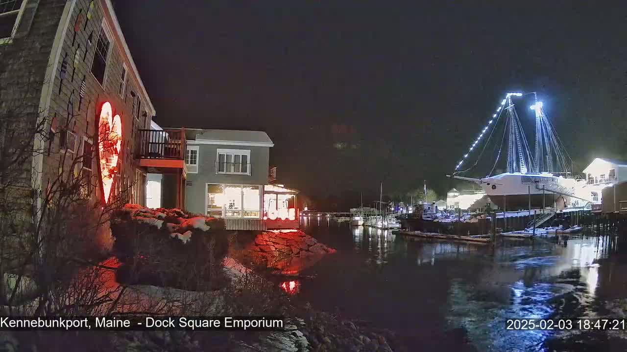 A nighttime view of a waterfront scene with snow on the ground, featuring a building illuminated by a large red heart-shaped light display, and a docked ship lit with white lights.