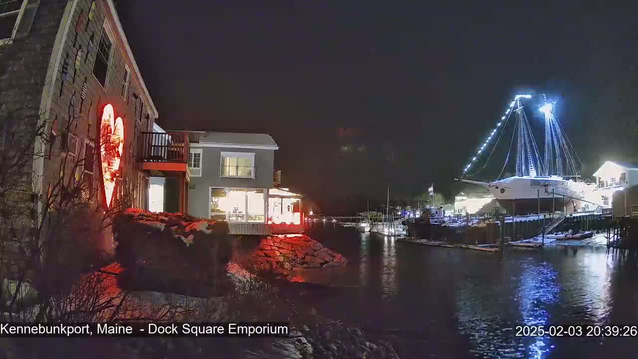 A nighttime waterfront scene shows a building illuminated with a large red heart, another building with lights, and a large ship docked, all lightly covered in snow.