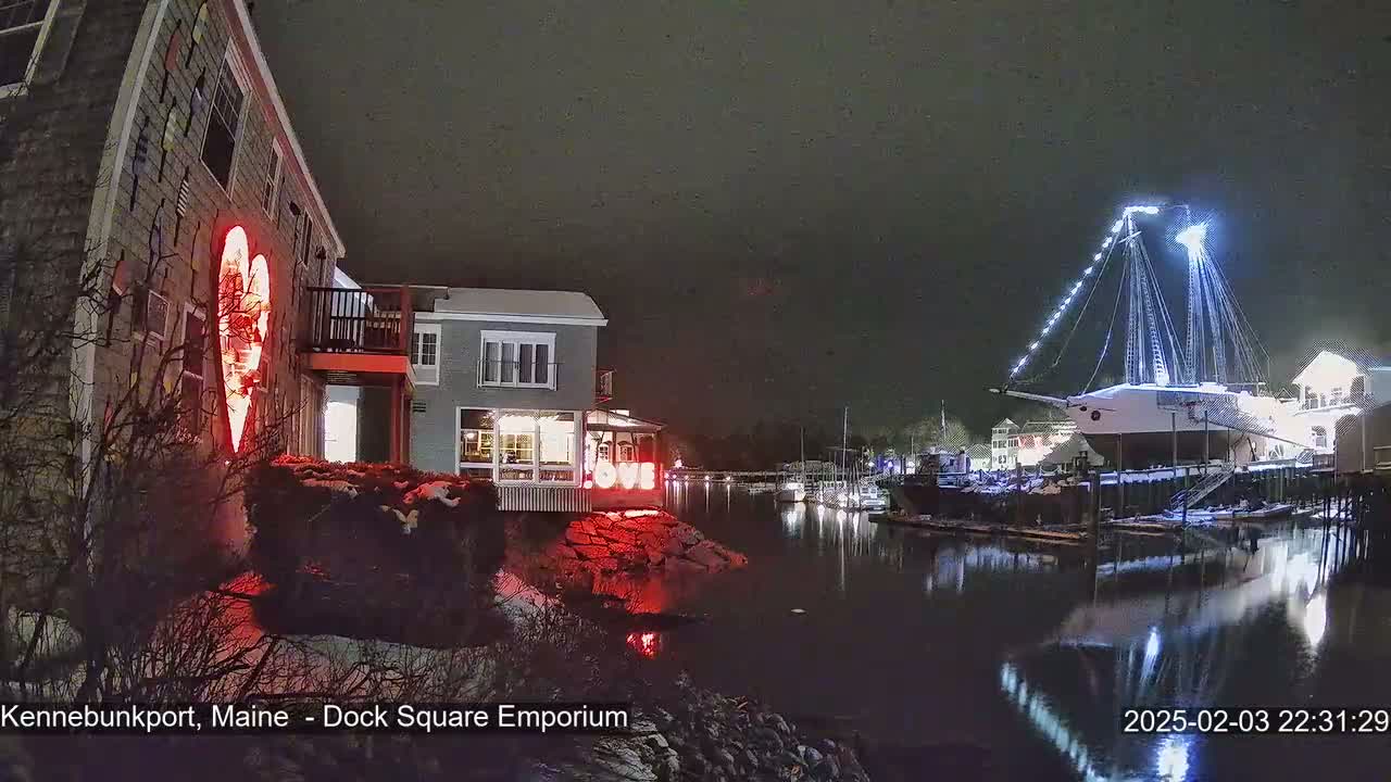 A nighttime view of a waterfront scene shows a building with a large illuminated heart on its side, another building across a waterway, and a large, illuminated ship docked nearby, with a dusting of snow on the ground.