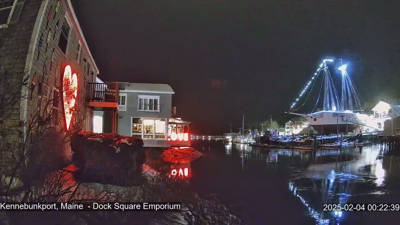 A nighttime waterfront scene shows a building with a lighted heart on its exterior and a large, illuminated sailboat docked nearby, with some snow on the ground.