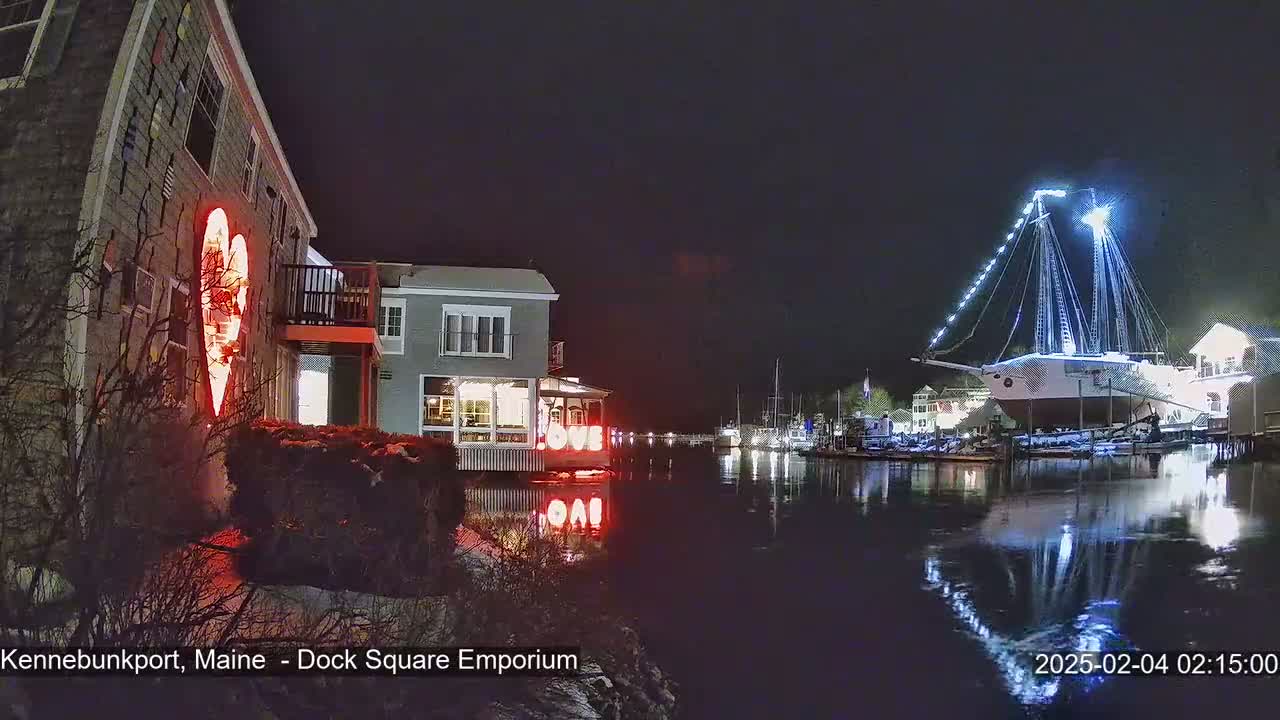 A nighttime harbor scene shows a building with a lit-up heart on its exterior, other buildings with lights, and a large, illuminated ship docked at the waterfront.