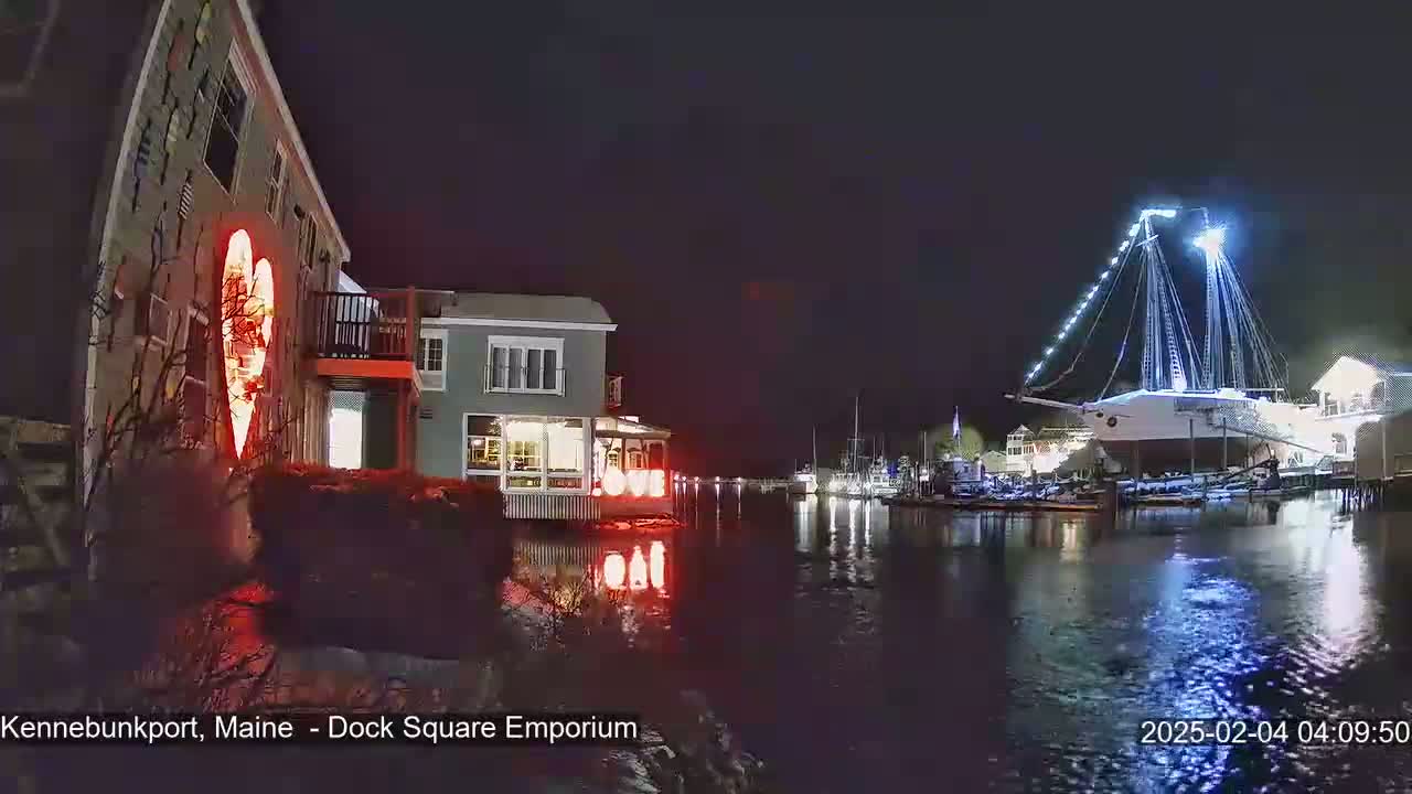 A nighttime waterfront scene shows a building illuminated with a large heart-shaped light display next to a calm body of water with a large, brightly lit ship docked in the distance.
