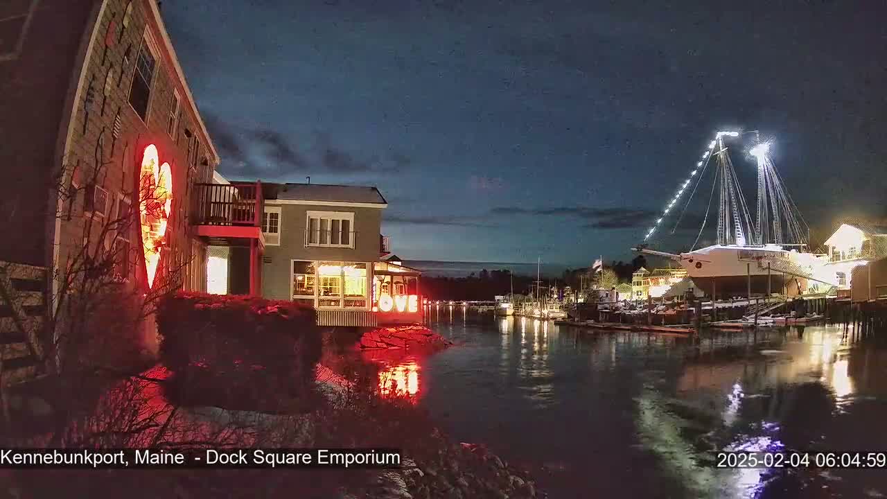A nighttime waterfront scene shows a building illuminated with a large red heart and the word "LOVE," a large sailing ship docked nearby, and calm water reflecting the lights.