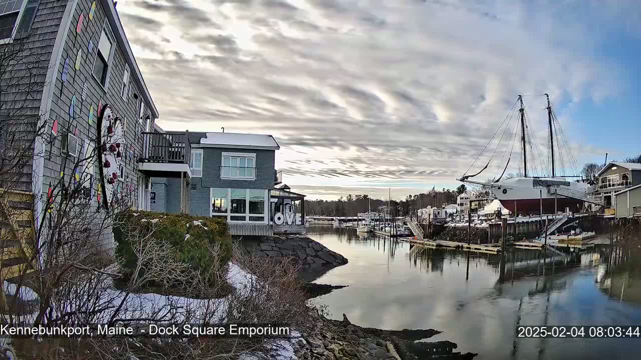 A waterfront scene with buildings, a large sailing ship docked, and a calm harbor under a partly cloudy sky.