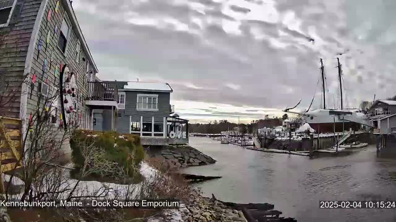 A waterfront scene on an overcast day shows a canal with docked sailboats and buildings with some snow on the ground.