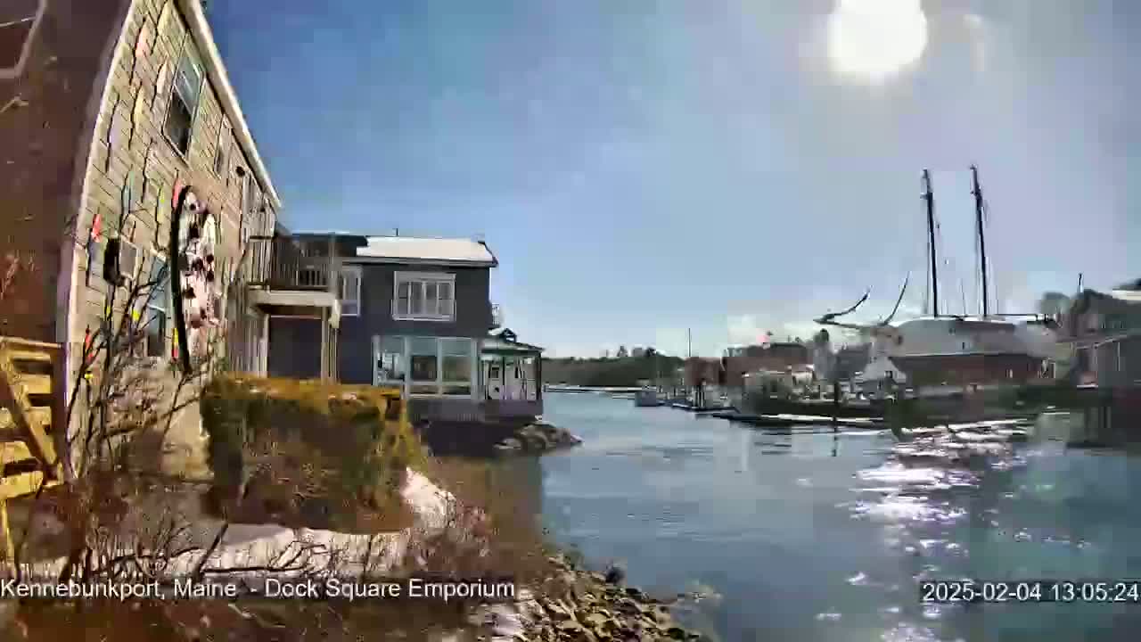 A sunny day shows a waterfront view of several buildings, some with snow on the ground, and a large sailing vessel docked in a calm harbor.