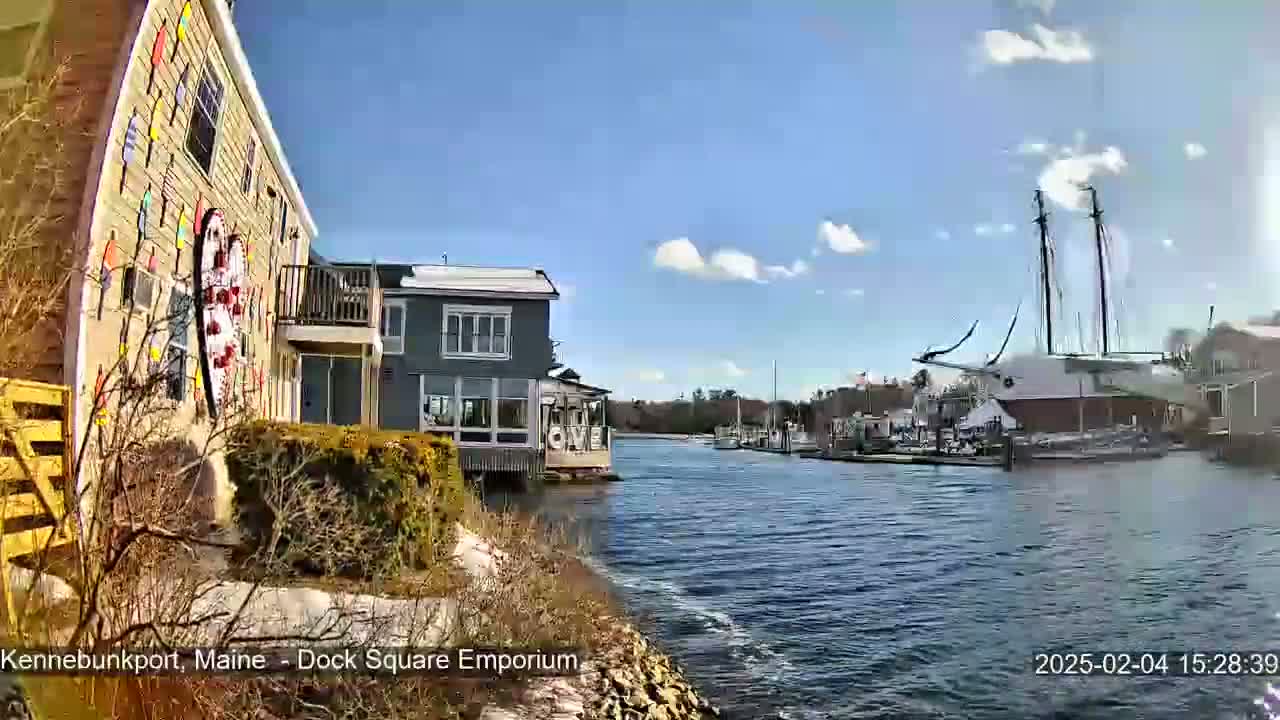 A waterfront scene on a sunny day with partly cloudy skies shows a calm body of water with docked boats, including a large sailing ship, and buildings along the shore, one of which displays a large heart-shaped decoration.