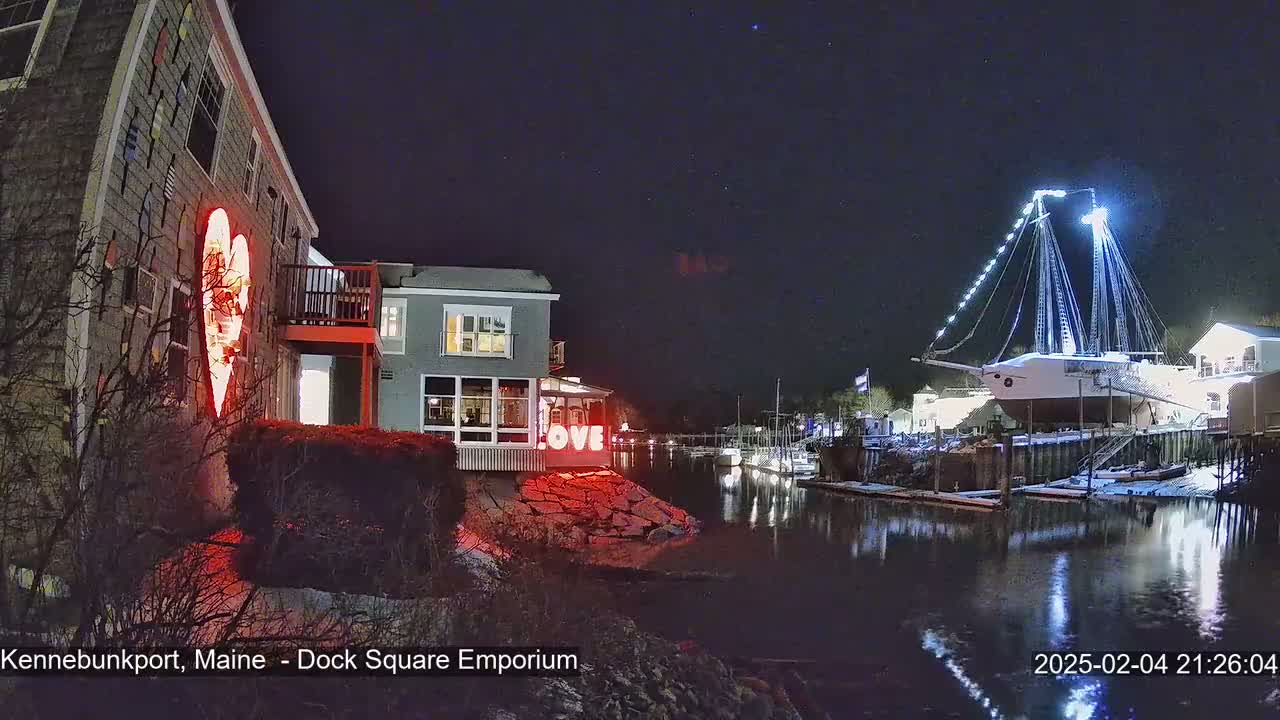 A nighttime waterfront scene shows a building with a large illuminated heart, another building with a lit "LOVE" sign, and a large ship illuminated with lights, all reflected in calm water under a dark, starry sky.