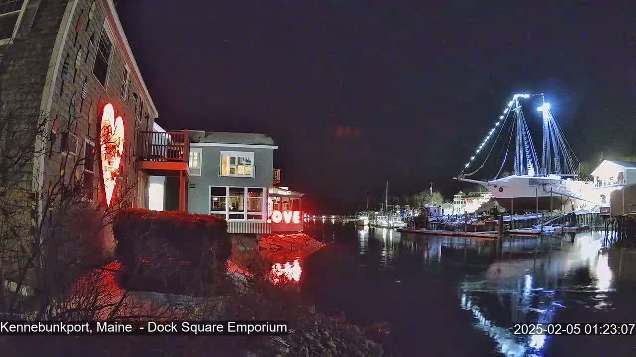 A nighttime scene of a waterfront area with buildings decorated with illuminated hearts and the word "LOVE," featuring a large sailboat lit up with lights, all under a dark sky.