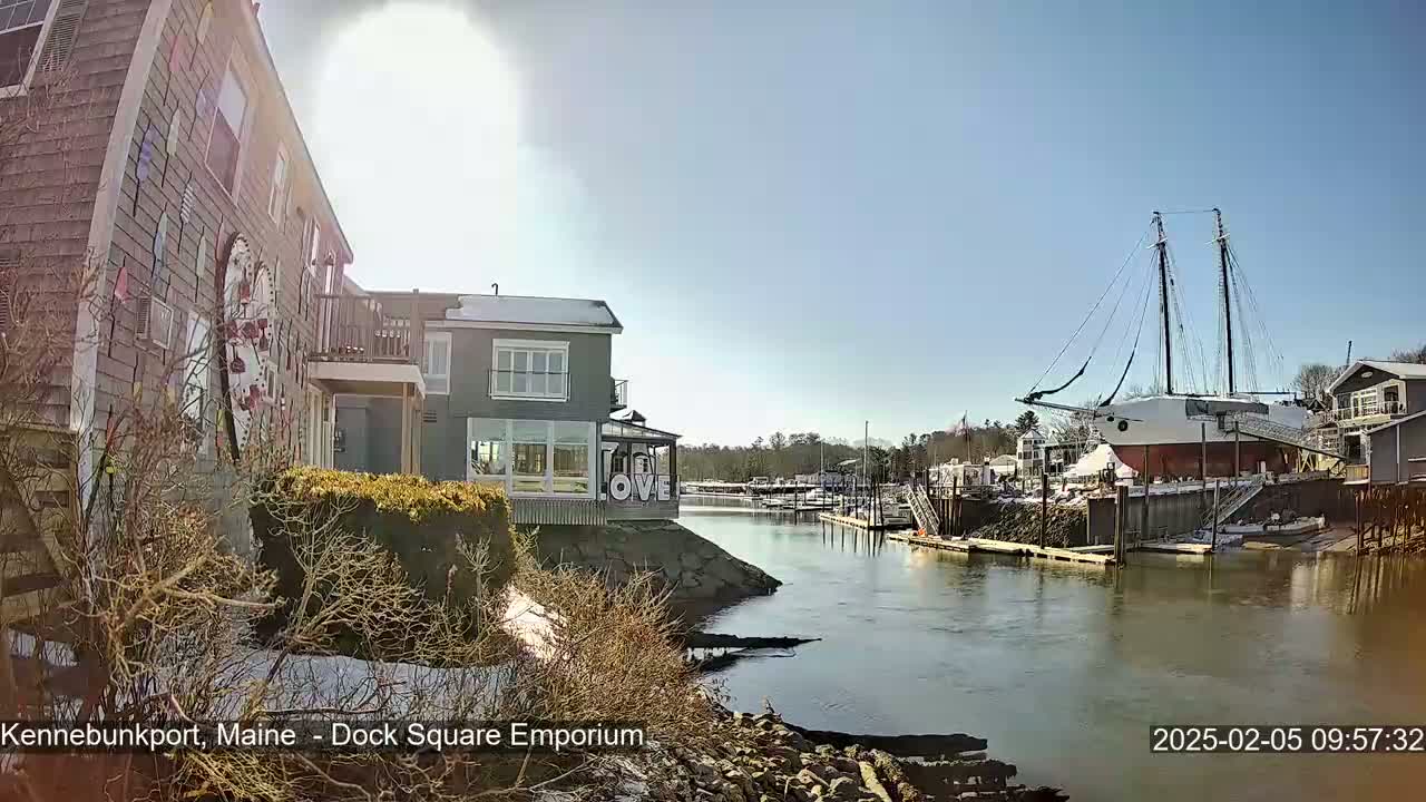 A sunny day reveals a waterfront scene with a large sailing ship docked near buildings, some with snow on the ground.