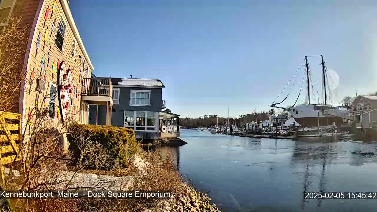 A waterfront view on a sunny day shows a building adorned with colorful buoys, other buildings, docked boats, and a large sailing ship in the harbor.