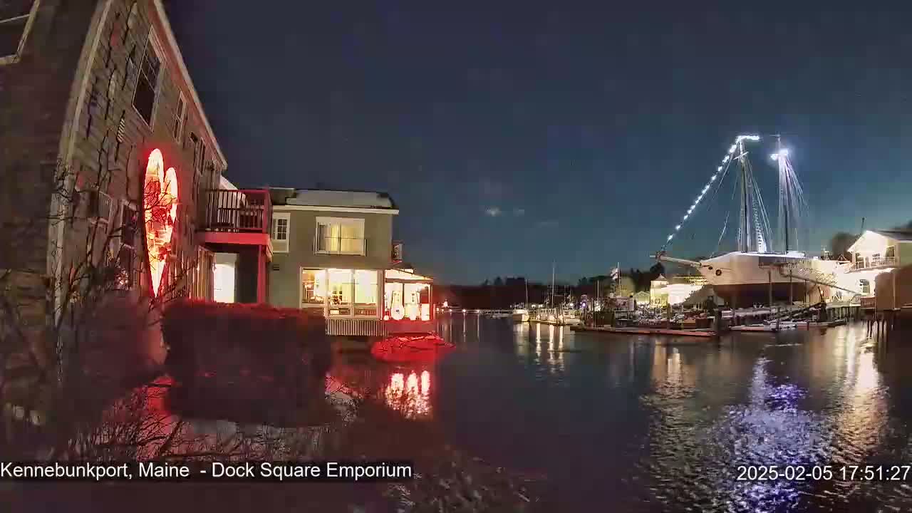 A nighttime view of a waterfront scene with a large sailboat docked, illuminated buildings, and a red heart-shaped light display on a building, all under a clear, dark sky.