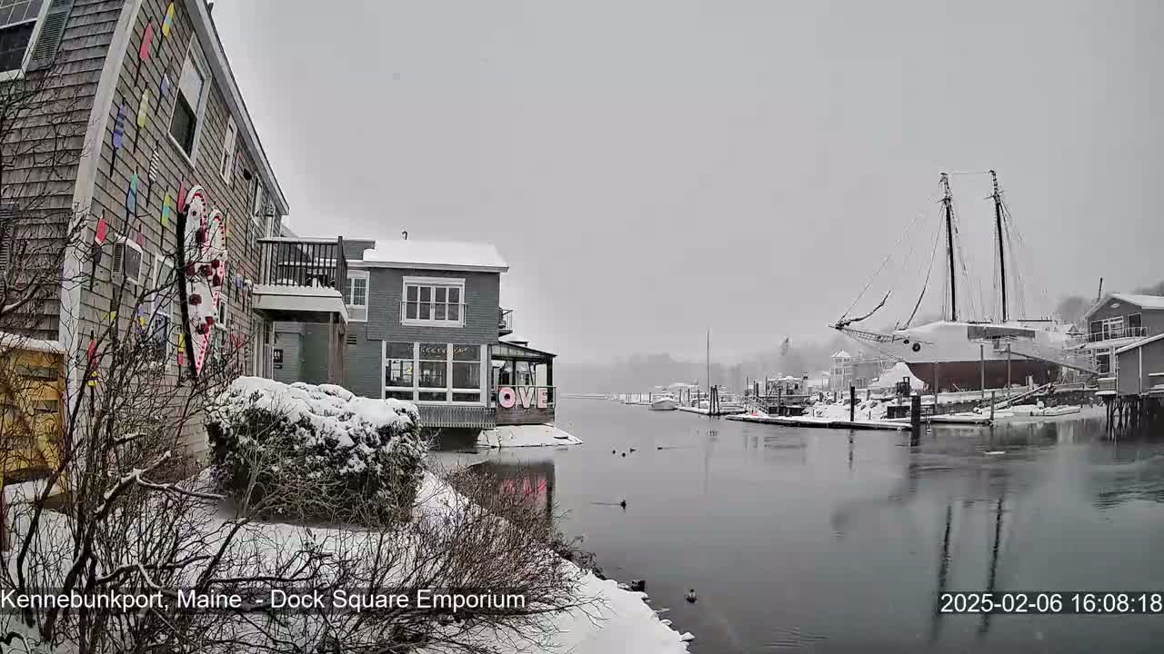 Snow falls on a waterfront scene featuring a building adorned with colorful nautical decorations, other buildings covered in snow, a large sailing vessel docked, and calm water with a few birds.
