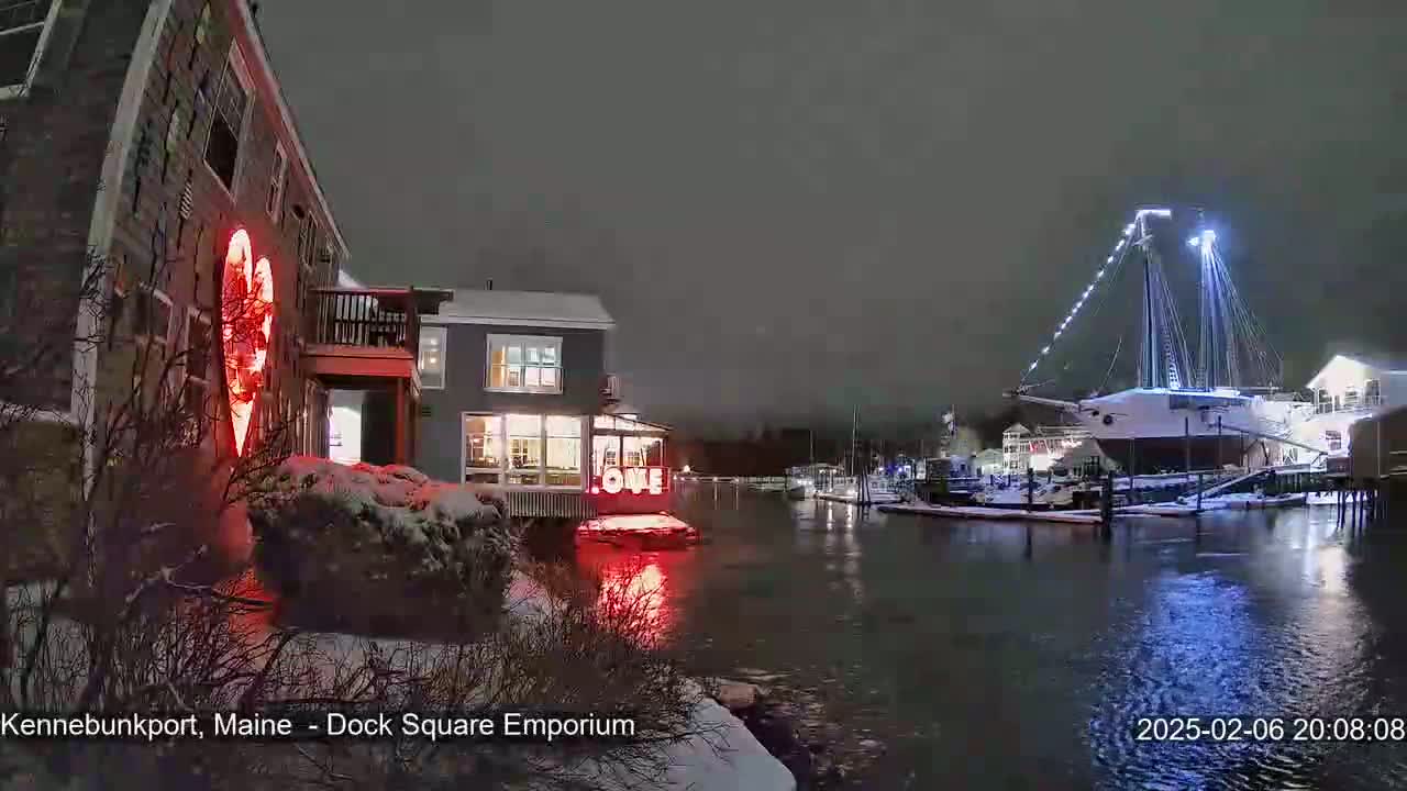 A nighttime scene shows a snow-covered harbor with a large sailboat, illuminated buildings, and a red heart-shaped light on a brick building.