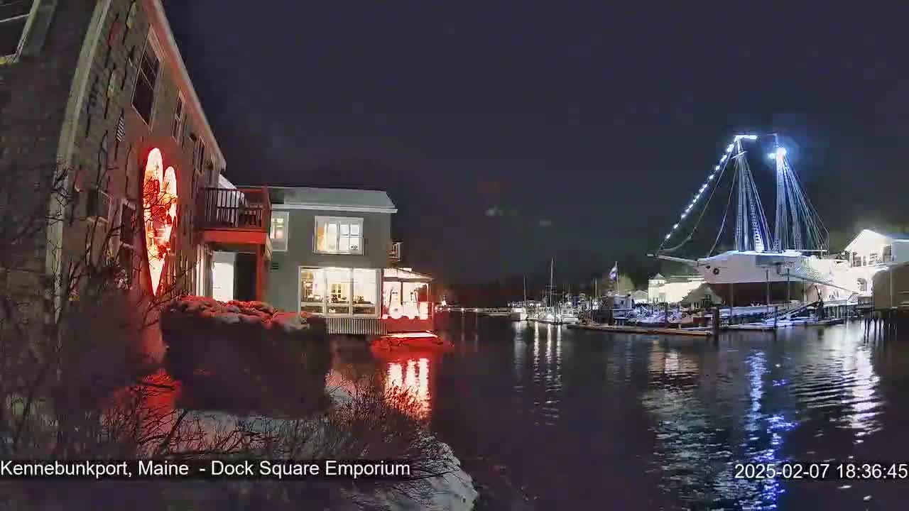 A nighttime waterfront scene shows a building with a large illuminated heart on its side, other buildings across the water, and a docked, lit-up sailing ship under a clear, dark sky; snow is on the ground.