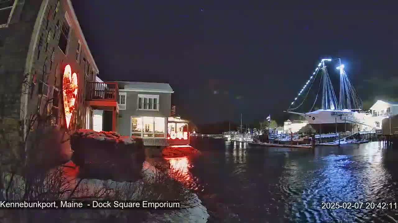 A nighttime waterfront scene shows a building with a large illuminated heart on its exterior, other buildings, boats, and a large sailing ship docked, all under a dark, clear sky with some snow on the ground.