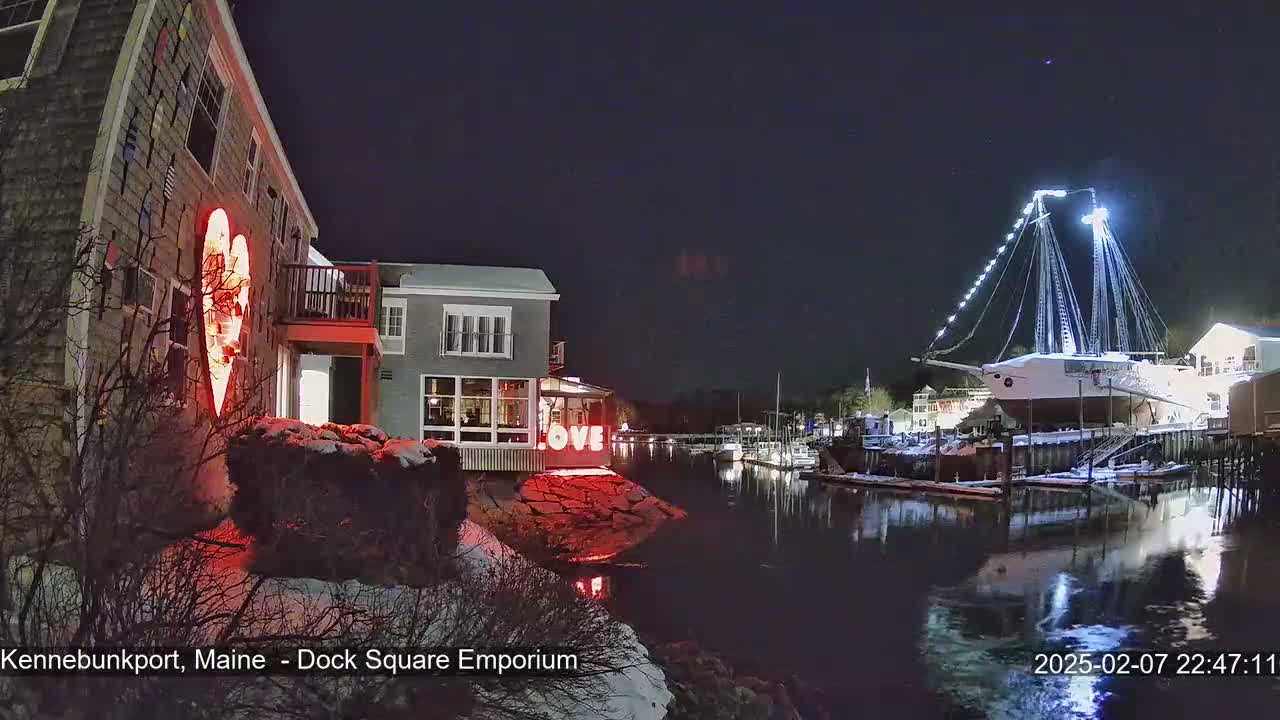 A nighttime waterfront scene shows a building with a large illuminated heart on its side, another building with lit windows, and a large sailboat docked, all under a clear, dark sky with some snow on the ground.