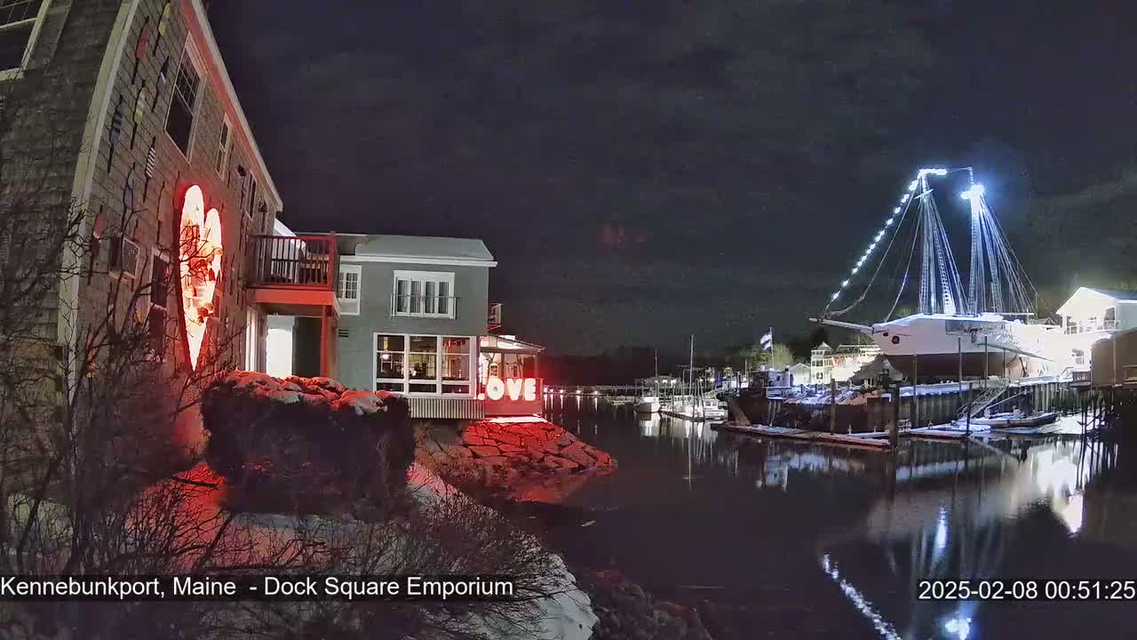 A nighttime harbor scene with snow on the ground shows a building illuminated with a large red heart, another building with the word "LOVE" displayed, and a large ship docked, all reflecting in the calm water under a dark, cloudy sky.