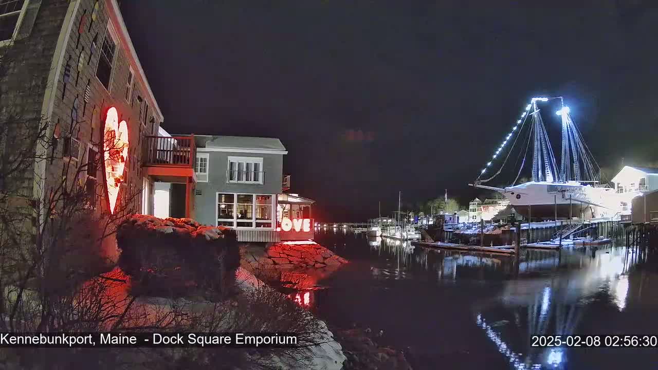 A snow-dusted waterfront scene at night shows a building lit with a red heart, another building with "LOVE" displayed, and a large sailboat docked, all reflected in the calm water.