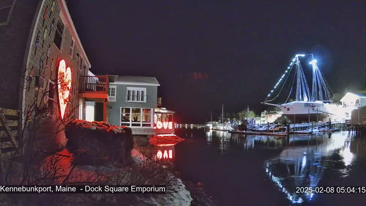 A nighttime waterfront scene shows a building with a large illuminated heart on its exterior, a brightly lit ship docked nearby, and a calm body of water reflecting the lights.