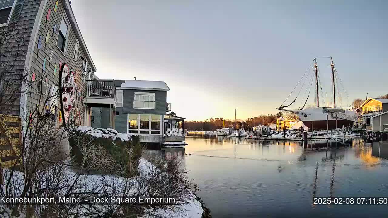 A snow-dusted waterfront scene at sunrise shows a calm harbor with docked boats, including a large sailing vessel, next to buildings with snow-covered grounds.