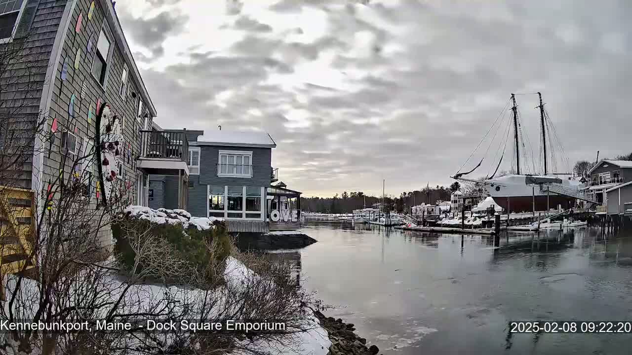 A snow-covered waterfront scene shows a colorful building, other buildings, a large sailboat, and a calm river under a cloudy sky.