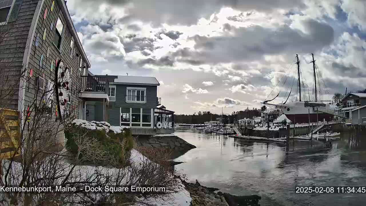 A partly cloudy day shows a snow-covered waterfront scene with buildings, a large sailboat, and a calm waterway.