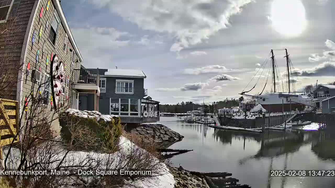 A partly cloudy day reveals snow-covered buildings and a docked sailboat near a waterway.