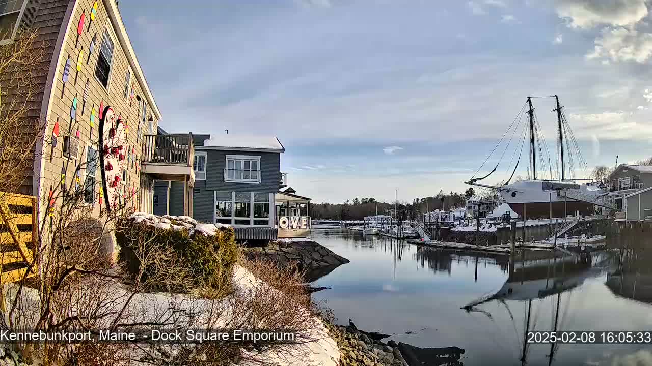 A snow-dusted waterfront scene shows a building decorated with colorful buoys and a large sailboat docked in a calm harbor under a partly cloudy sky.