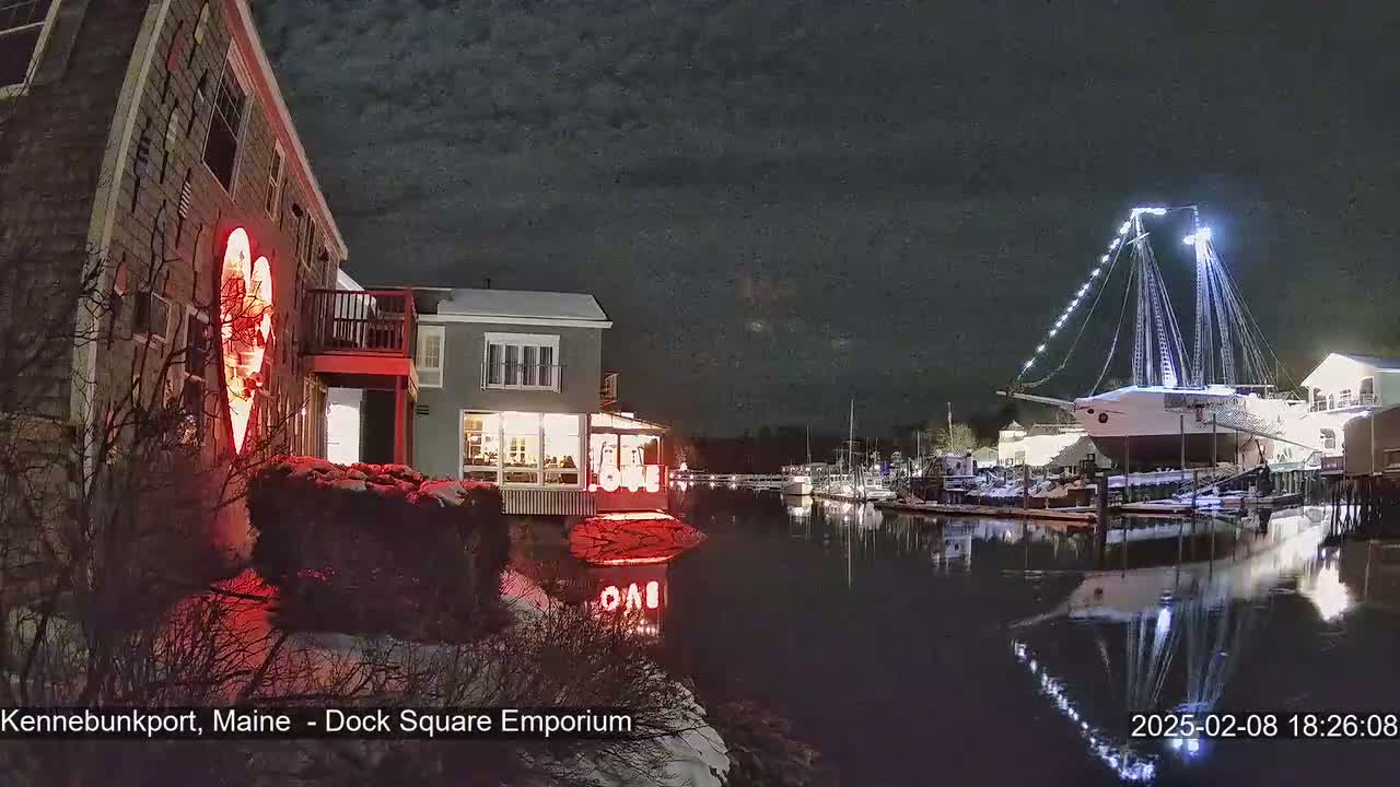 A nighttime waterfront scene shows a building with a large illuminated heart on its side, other buildings, a docked ship with lights, and calm water reflecting the lights.