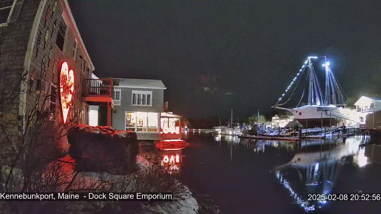 A nighttime harbor scene shows a building with a large illuminated heart on its exterior, a docked sailboat with lights, and snow on the ground.