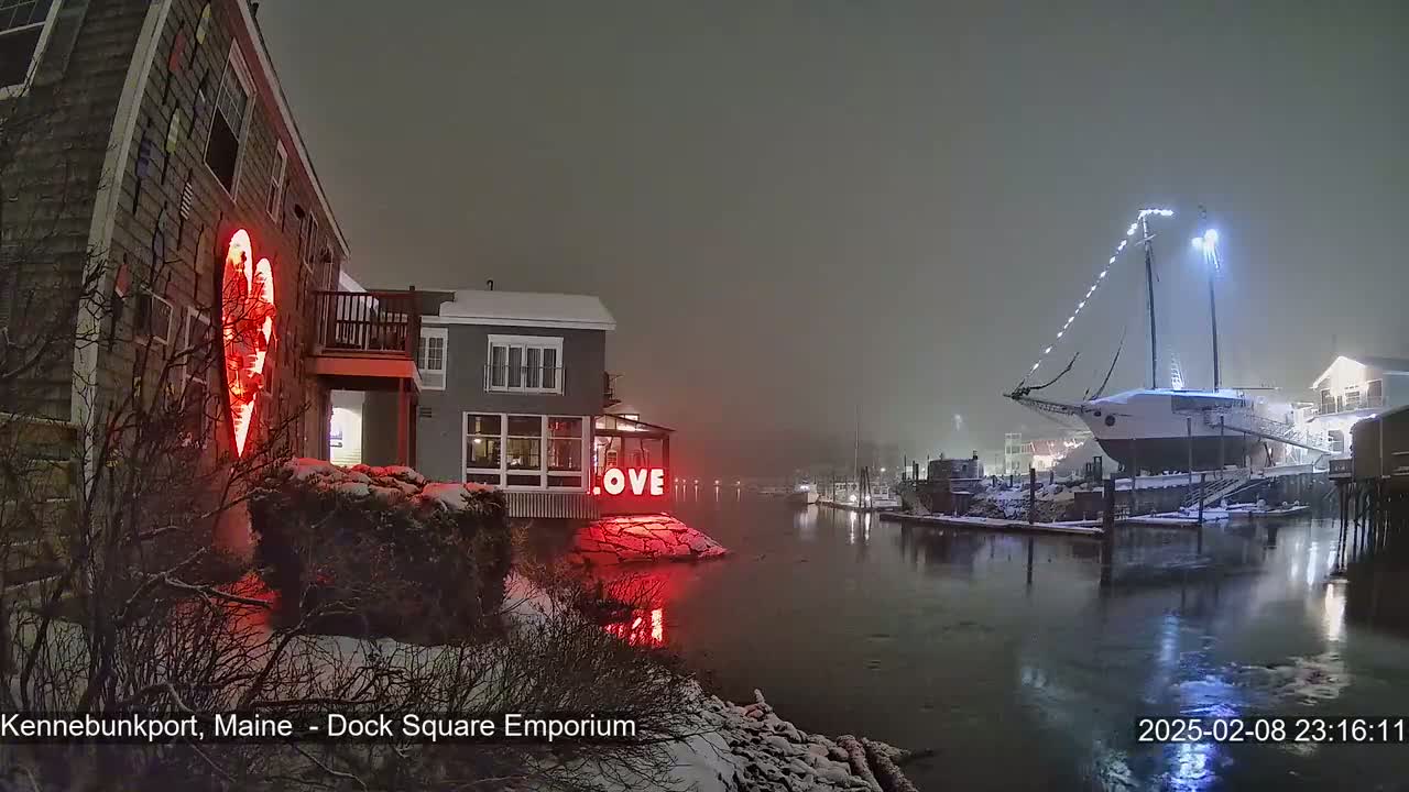 A snow-covered waterfront scene at night shows a building with a large red heart illuminated on its exterior, a lit-up "LOVE" sign, and a large sailboat docked nearby.