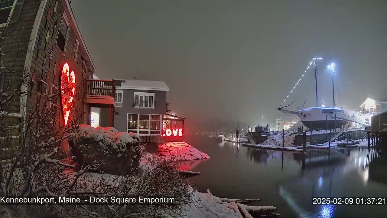 A snow-covered waterfront scene at night features a building with a red illuminated heart and the word "LOVE" in lights, alongside a docked sailboat under a hazy sky.