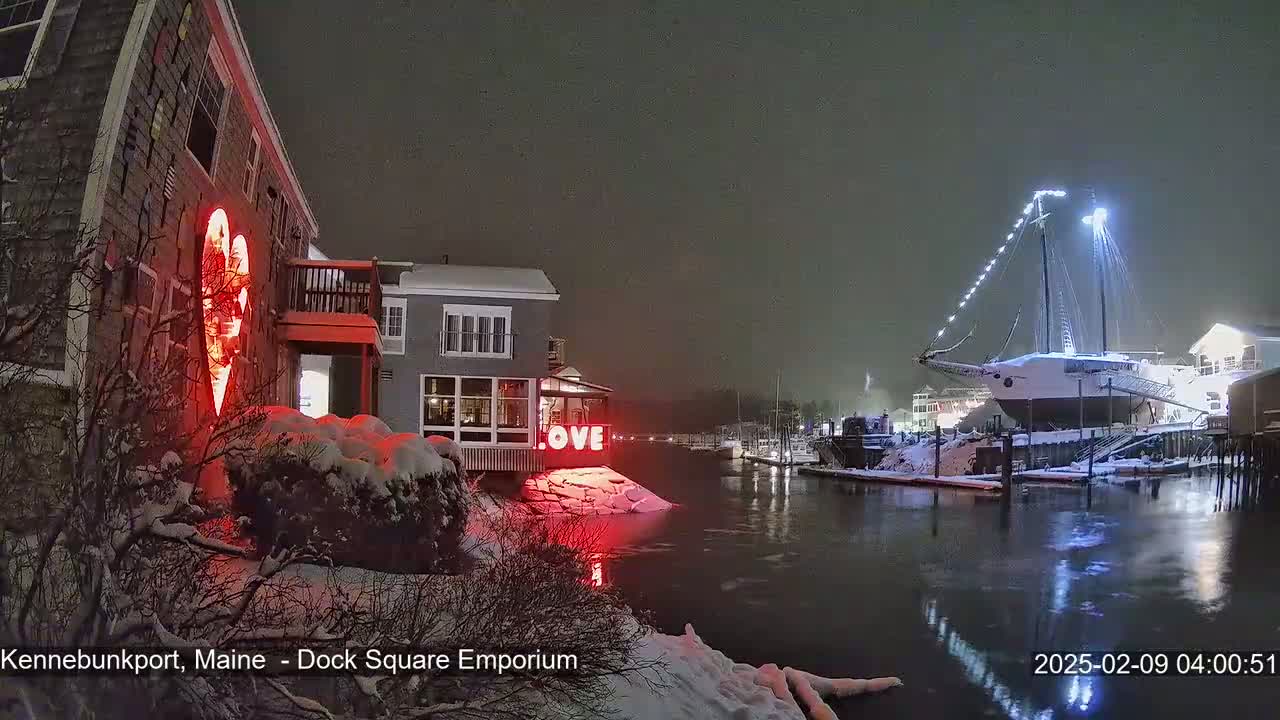 A snow-covered waterfront scene at night shows a building with a red illuminated heart, a sign that says "LOVE," and a large sailboat docked nearby.