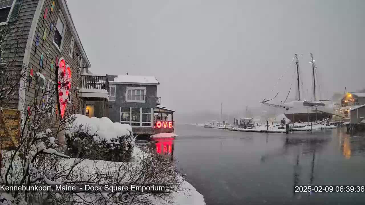 A snow-covered waterfront scene shows a building with a large illuminated heart and the word "LOVE,"  a docked sailboat, and calm water under a heavy snowfall.