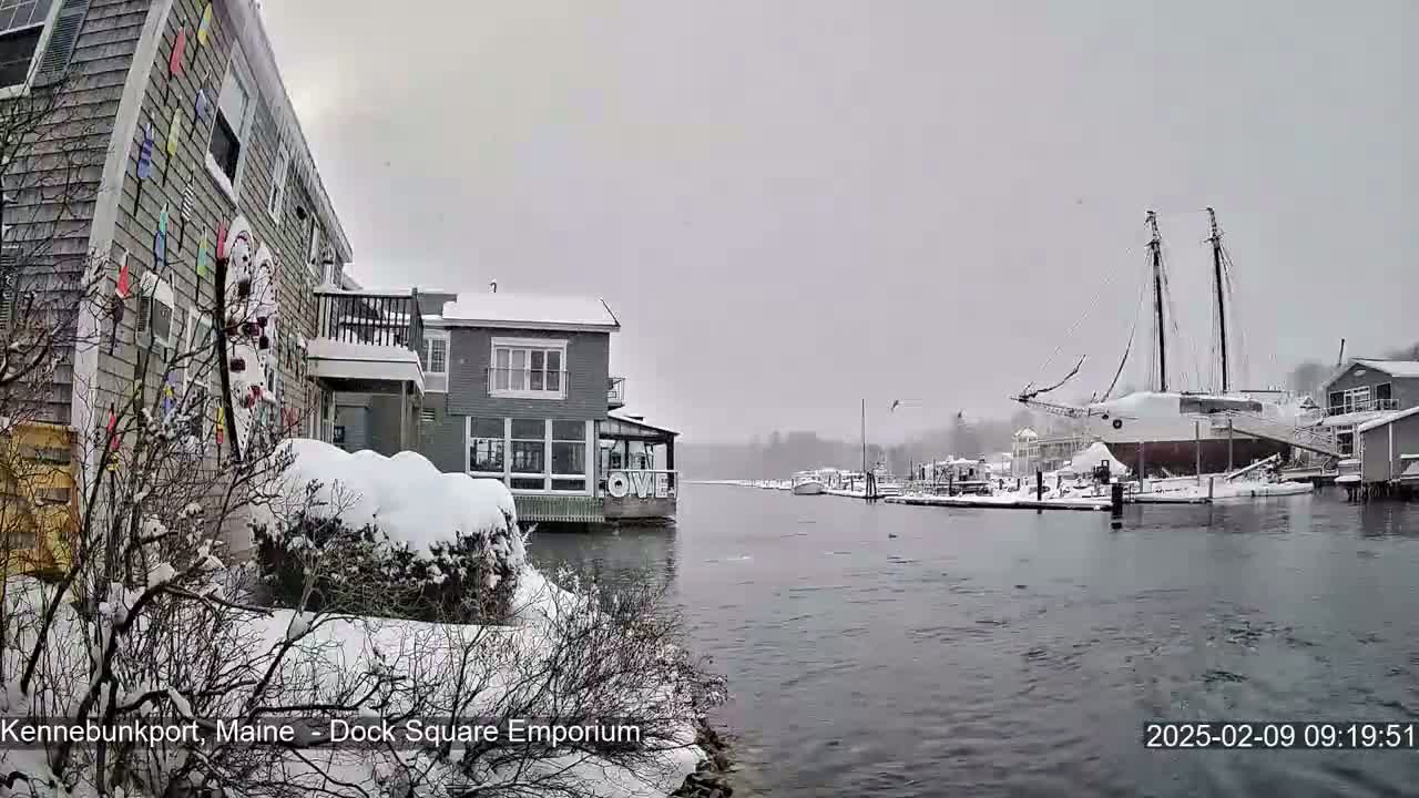 A snow-covered waterfront scene shows buildings and a large sailboat docked in the harbor during a light snowfall.
