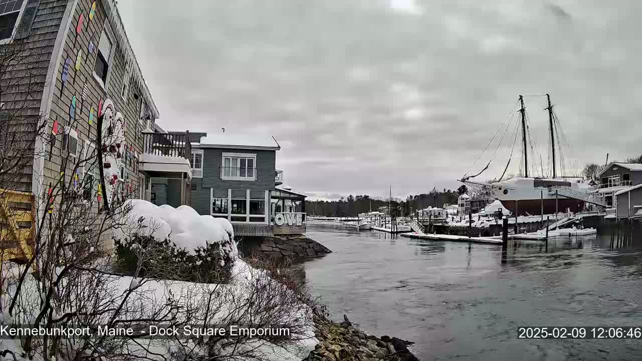 A snow-covered waterfront scene shows a harbor with a large sailboat and several buildings, under an overcast sky.