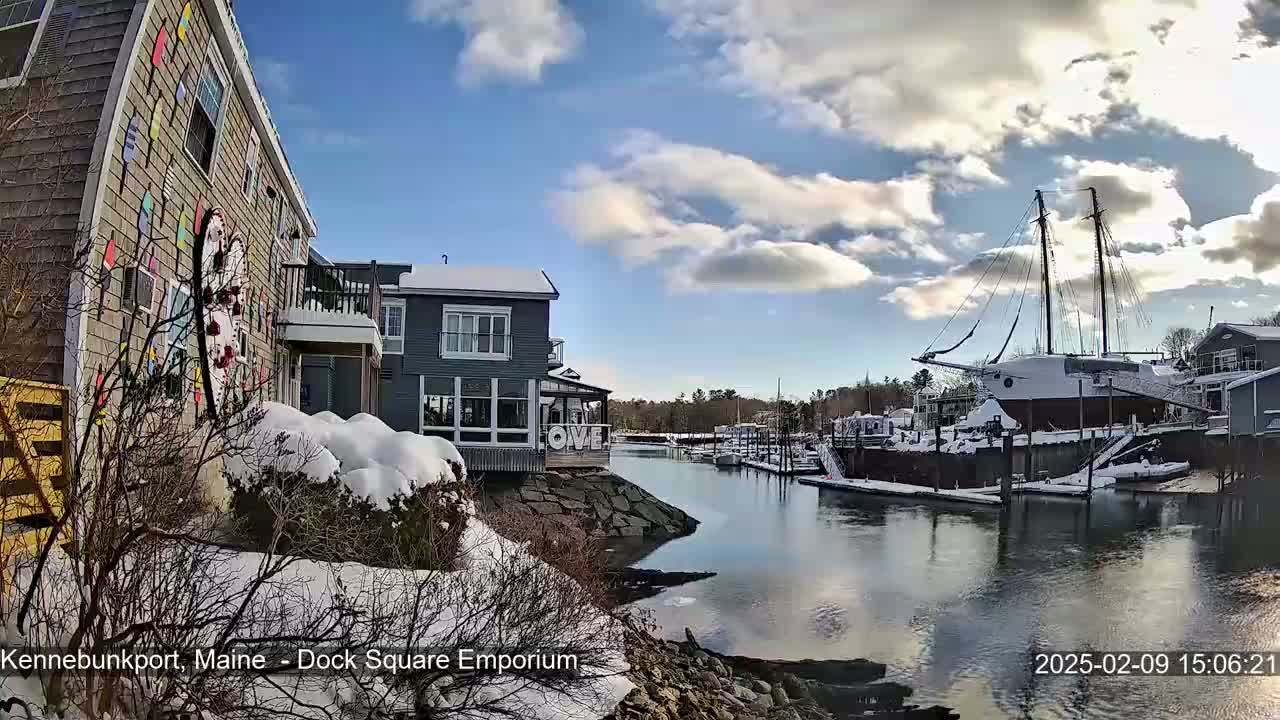 A snow-covered waterfront scene shows buildings, a docked sailboat, and a calm waterway under a partly cloudy sky.