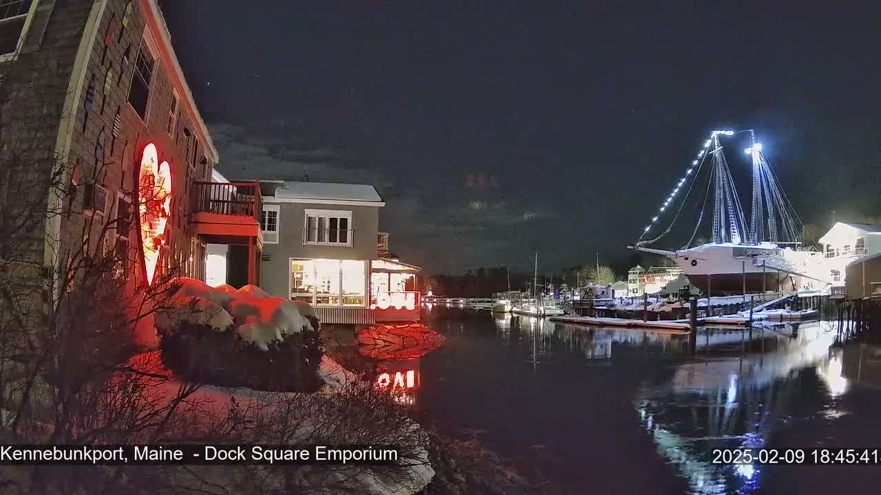 A snow-covered waterfront scene at night shows a building with a large illuminated heart, other buildings, a docked sailboat, and calm water reflecting the lights.