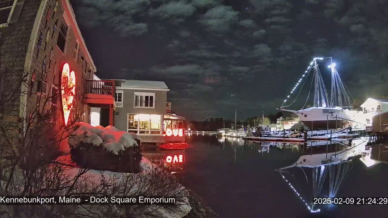 A nighttime scene of a snow-covered waterfront with buildings illuminated, a large sailboat at dock, and a heart-shaped light display on one building.