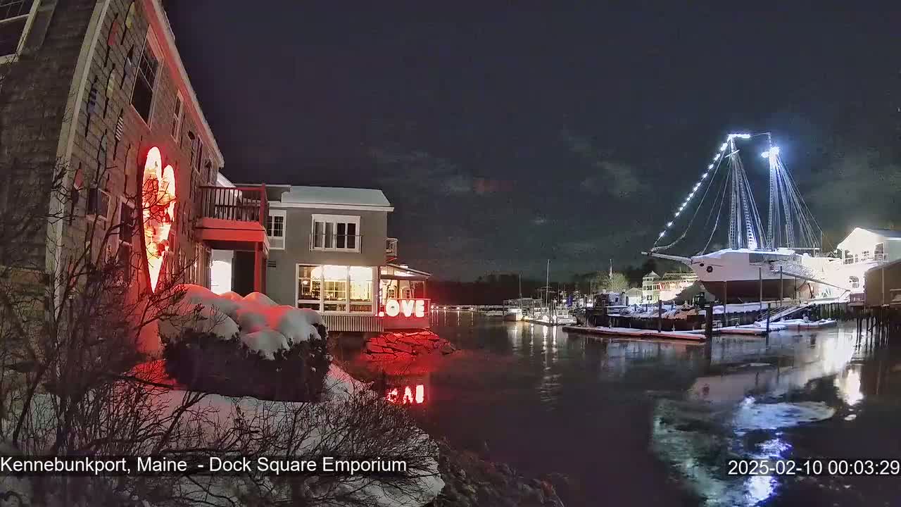 A snow-covered waterfront scene at night shows a building with illuminated heart decorations, a large sailing ship docked, and calm water reflecting lights.