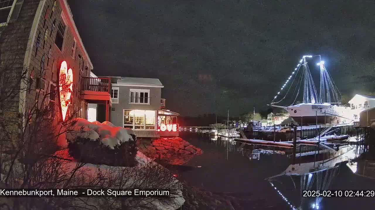 A snow-covered waterfront scene at night shows illuminated buildings, one with a large heart-shaped light display, and a large sailing ship docked, its reflection visible in the calm water.