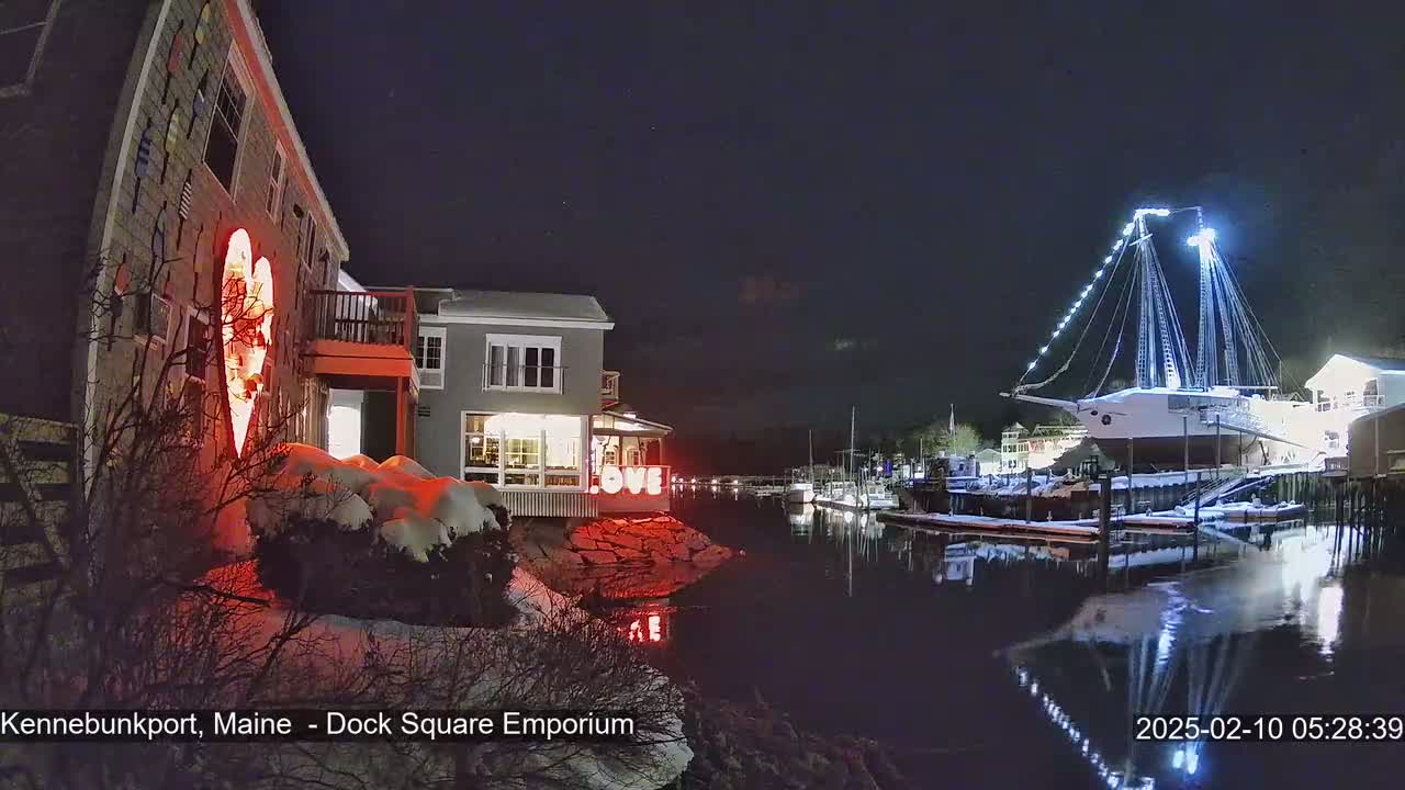 A snow-covered waterfront scene at night shows a building with a red heart-shaped light and the word "LOVE" illuminated, and a large, illuminated sailing ship docked nearby.