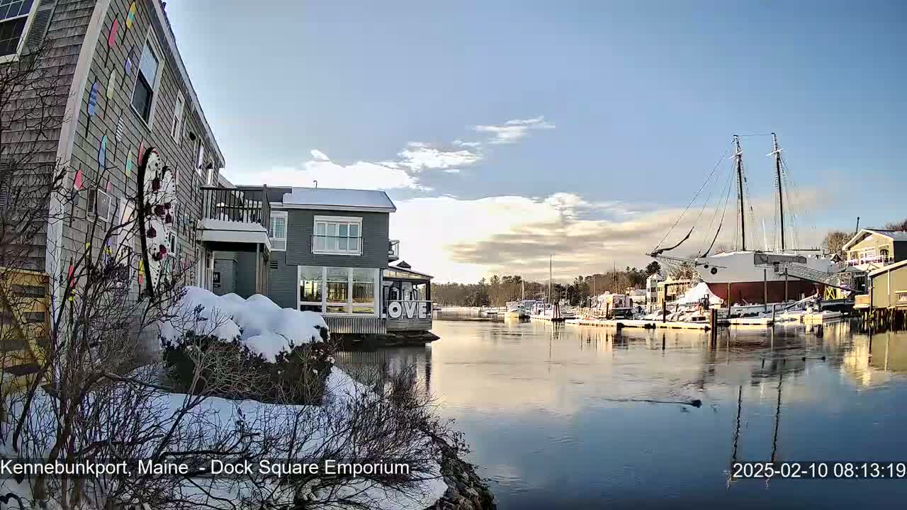 A snow-covered waterfront scene shows buildings, boats docked in calm water under a partly cloudy, sunny sky.