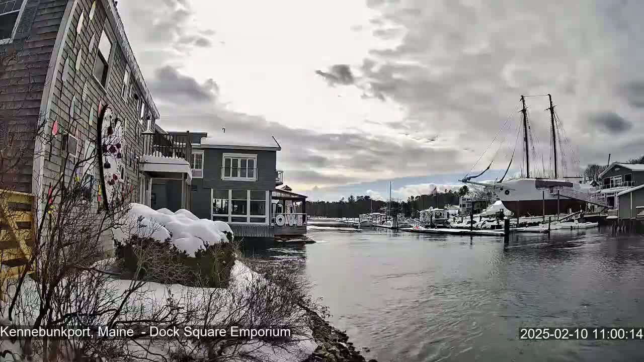 A snow-covered waterfront scene shows several buildings and a large sailboat docked in a calm waterway under a cloudy sky.