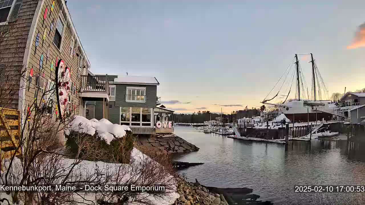 A snow-covered waterfront scene at sunset shows colorful buildings, a large sailboat docked, and several smaller boats in a calm harbor.