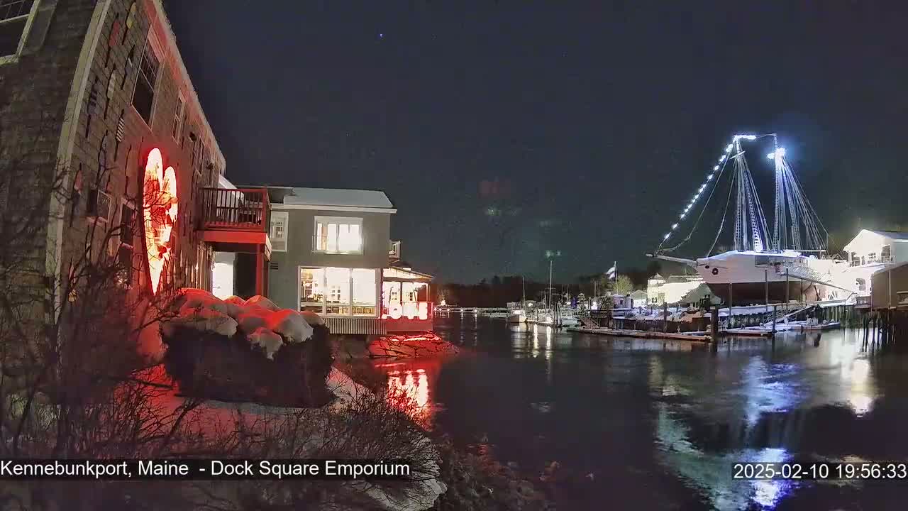 A nighttime waterfront scene shows a building with a large illuminated heart on its side, another building with lit windows, a large ship docked at a pier, and snow on the ground, all under a clear, dark sky.