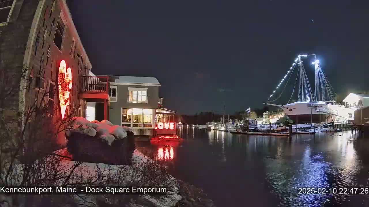 A snow-covered waterfront scene at night shows a large illuminated heart on a building, the word "LOVE" in lights nearby, and a large ship docked in the harbor.