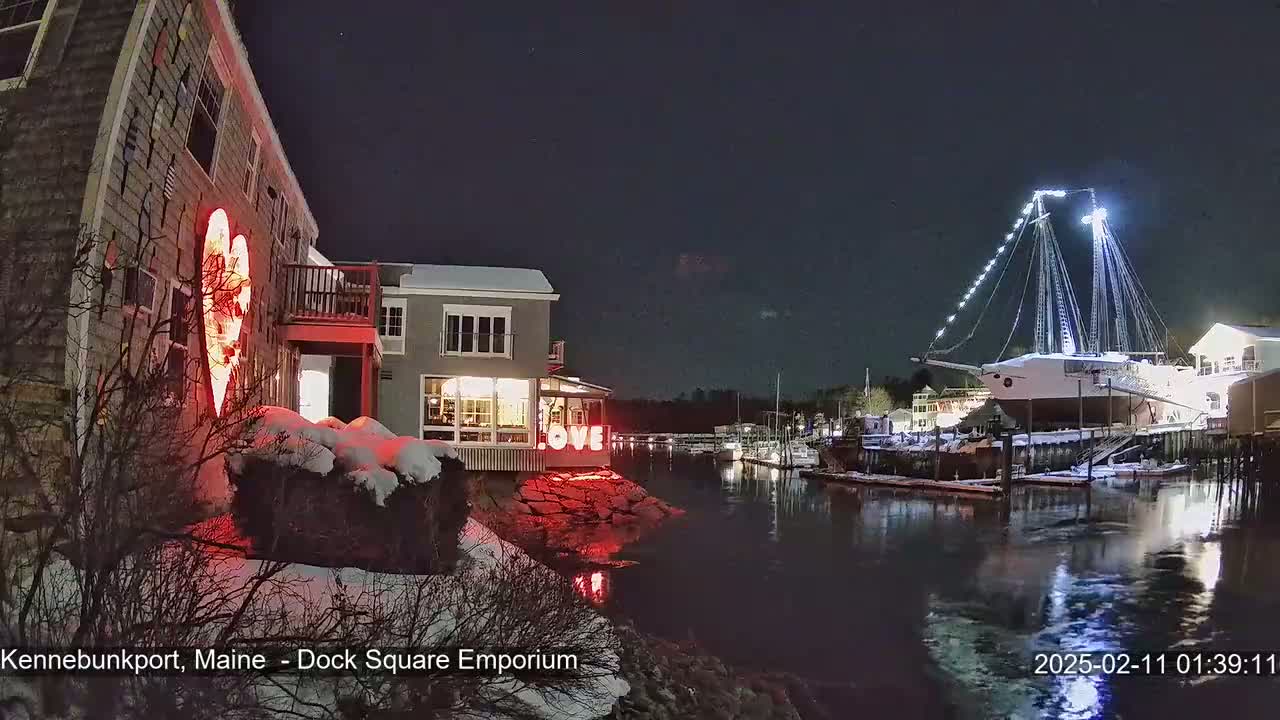 A snow-dusted waterfront scene at night shows a building with a large illuminated heart on its exterior, another building with illuminated letters spelling "LOVE," and a large, illuminated ship docked nearby.